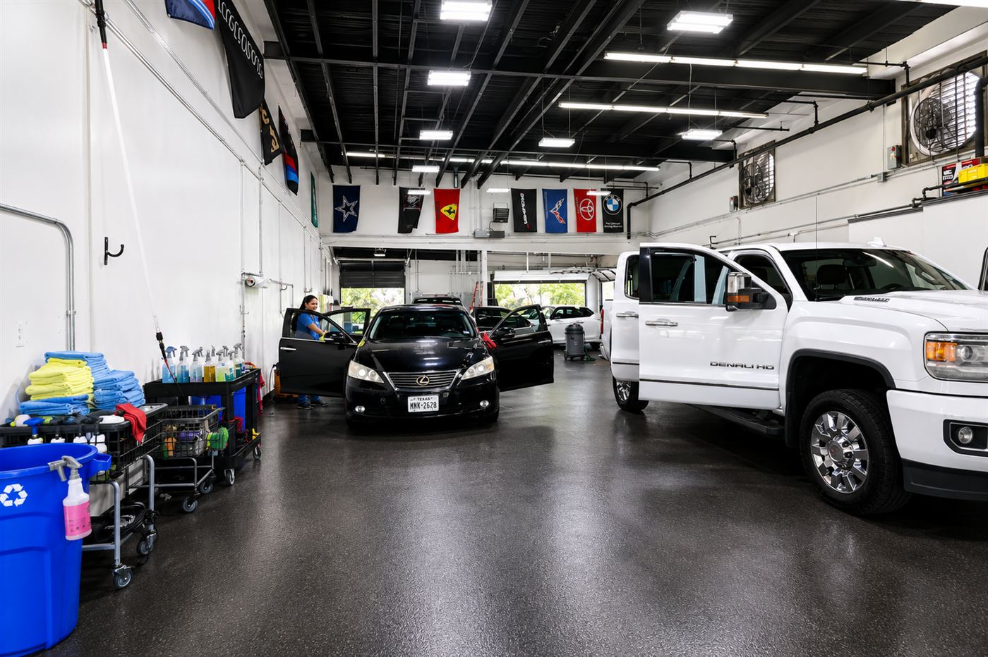 Bright, clean indoor detail bay with a white pickup and dark sedan being worked on