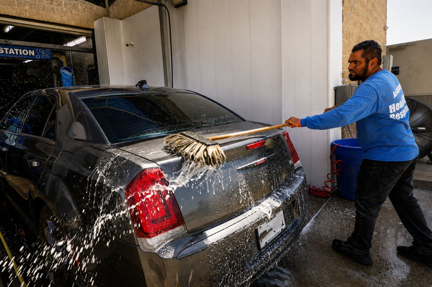 Crew member hand-washing the rear of a dark sedan with a soft mitt as water sprays